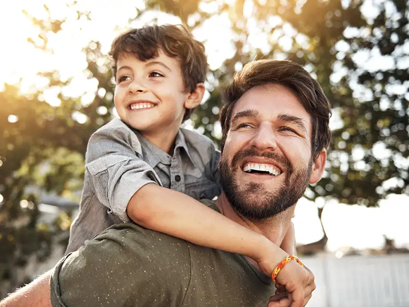 father smiling with son riding on his back outdoors near trees